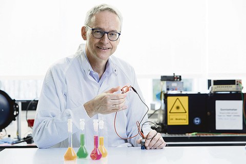 Prof. Leo sitzt an einem Labortisch. Im Hintergrund sind verschiedene Geräte zu sehen. Er hält eine Leuchtdiode in der Hand.