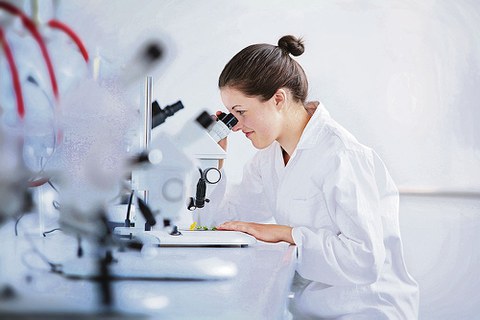 Eine Wissenschaftlerin in weißem Kittel sitzt an einem Tisch. Sie schaut in ein Mikroskop.