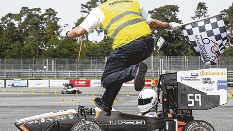 Ein Mann mit gelber Weste und schwarz-weiß-karierter Flagge in der Hand springt über einen Sportwagen. Darin sitzt ein Fahrer/eine Fahrerin.
