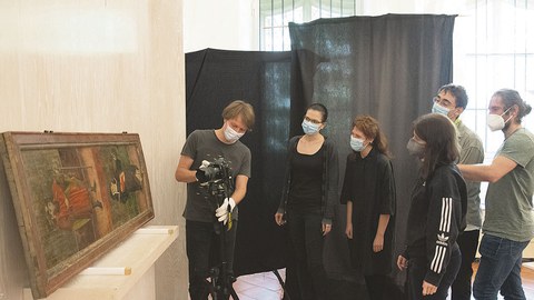 Ein Bildnis ist auf einem Tisch aufgestellt und an die Wand gelehnt. Davor stehen fünf Personen. Eine Person betreut einen Fotoapparat, der auf einem Stativ vor dem Tisch steht.