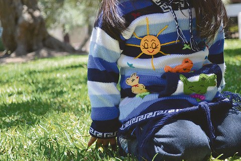 Auf dem Foto kniet ein Kleinkind auf einer Wiese. Zu sehen ist nur der Körper, kein Gesicht. Der blau-gestreifte Pullover ist bestickt mit fröhlichen Motiven.
