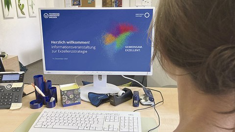 Auf einem Schreibtisch steht ein Computer-Monitor. Auf diesem ist das Startbild der Informationsveranstaltung zu sehen.