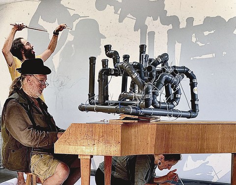 Prof. Schmidt sitzt an einem Flügel. Auf dem Flügel steht ein Objekt aus dunklen Kunststoffrohren. Unter dem Flügel ist eine Frau zu sehen, daneben steht ein dirigierender Mann.