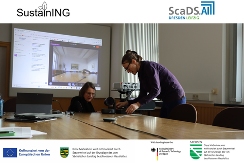 In the foreground is a person leaning over a table and working on a laptop. Another person is sitting next to them, looking at the screen. In the background, a large monitor is visible, showing a video conference with a camera feed of an empty meeting room. The room is bright and has large windows with vertical blinds. Various technical devices are on the table. The ScaDS.AI and SustainING logos are visible at the top of the image. Funding acknowledgments are visible at the bottom of the image. 