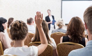 Foto von Zuhörern in einem Seminar. Die Zuhörer sind mit dem Rücken dem Betrachter zugewandt und einer von ihnen meldet sich.