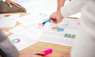 Das Foto zeigt mehrere Personen, die um einen Tisch stehen. Auf dem Tisch liegen zahlreiche ausgedruckte Statistiken und Diagramme.