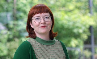 Foto von einer jungen Frau mit roten Haaren und eine Brille in grünem T-Shirt und grüner Jacke. Im Hintergrund verschwommene Bäume.