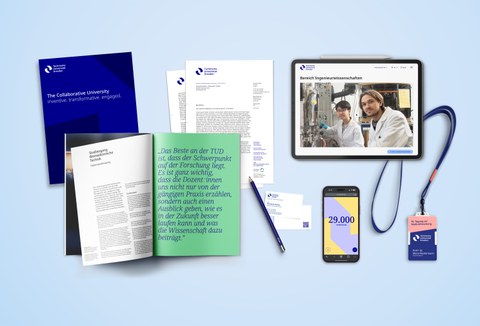 Various branding elements of TU Dresden including a dark blue booklet with white text, an open magazine with green and white text, a letterhead, a pen, a smartphone displaying '29,000', a tablet showing a webpage, and an ID badge with a blue lanyard.