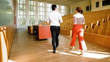 Foto: Zwei Dozenten gehen in einem hellen Hörsaal zum Sprechpult, sie sind von hinten zu sehen. Der Hörsaal hat große Fenster und eine Holzbestuhlung.