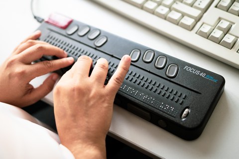 Photo of a Focus 40 Blue Braille display in front of a computer keyboard. Two hands touch the pins on the Braille display.