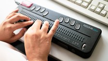 Photo of a Focus 40 Blue Braille display in front of a computer keyboard. Two hands touch the pins on the Braille display.