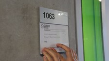 Photo of two hands touching a transparent Braille label on a door sign. The doorplate shows the room number 1063 and there is a logo of the TU Dresden on top. Underneath is the german writing "Institut für Angewandte Informatik, Professur für Mensch-Computer Interaktion, LAS".
