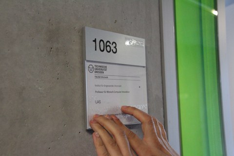 Photo of two hands touching a transparent Braille label on a door sign. The doorplate shows the room number 1063 and there is a logo of the TU Dresden on top. Underneath is the german writing "Institut für Angewandte Informatik, Professur für Mensch-Computer Interaktion, LAS".