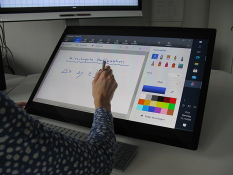 Photo of a switched on Microsoft Studio. The display shows a drawing surface with setting options on the right and a drawing surface on the left. In front of the computer sits a person who draws on the drawing surface.