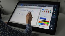 Photo of a switched on Microsoft Studio. The display shows a drawing surface with setting options on the right and a drawing surface on the left. In front of the computer sits a person who draws on the drawing surface.