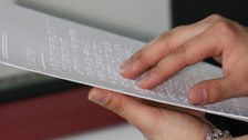 Photo of a Braille printout. One hand is holding the paper, the other is feeling the Braille.