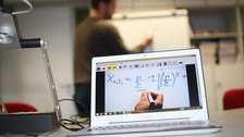 Photo of an exemplary teaching situation. In the foreground there is a laptop on the right and a panel camera on the left. In the background you can see a person drawing on a flipchart. On the screen of the laptop you see a close-up of a white surface with a drawing hand and a formula above it.