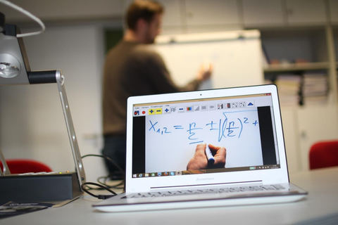 Photo of an exemplary teaching situation. In the foreground there is a laptop on the right and a panel camera on the left. In the background you can see a person drawing on a flipchart. On the screen of the laptop you see a close-up of a white surface with a drawing hand and a formula above it.