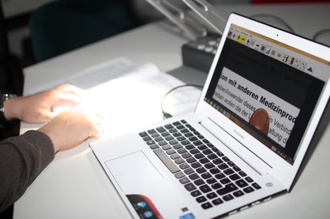 Photo of a laptop and a panel camera behind it. Below the camera is a book on which a person has placed two hands. On the screen you can see the close-up of a book page. Under a line of text there is a human finger.