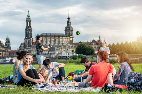Foto: Gruppe von Studierenden an der Elbe. Sie spielen Volleyball und sitzen plaudernd auf der Wiese.