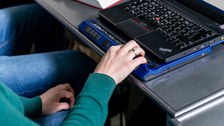 Foto einer Person, deren Arm zu sehen ist. Die Person sitzt an einem Laptop und ihre Hand liegt auf einer Braillezeile. 