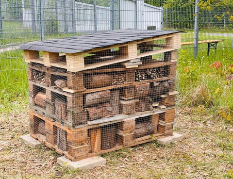 Nahaufnahme eines Insektenhotels aus 6 übereinander gestapelten Europaletten, welches in den Zwischenräumen mit zahlreichen angeborten Holzstücken gefüllt wurde. Eine angeschrägte mit Dachpappe bedeckte Palette schützt das Hotel vor Regen.  