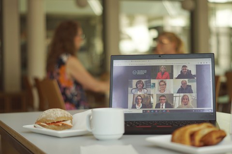 Laptop und Kaffeetassen auf einem Tisch in der Mensa, im Hintergrund sprechen zwei Menschen miteinander