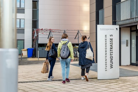 Das Foto zeigt drei Studierende, die den Eingang eines Studentenwohnheims ansteuern. Sie haben Rucksäcke und Tüten dabei.