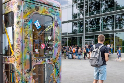 Das Foto zeigt eine bemalte Telefonzelle. Sie steht vor dem Hörsaalzentrum der TU Dresden. In der Telefonzelle befinden sich viele Bücher. Auf den Treppen vor dem Hörsaalzentrum sitzen viele Studierende.