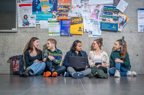 Fünf junge Menschen sitzen lachend auf dem Boden vor einer Pinnwand voller Aushänge und unterhalten sich miteinander.