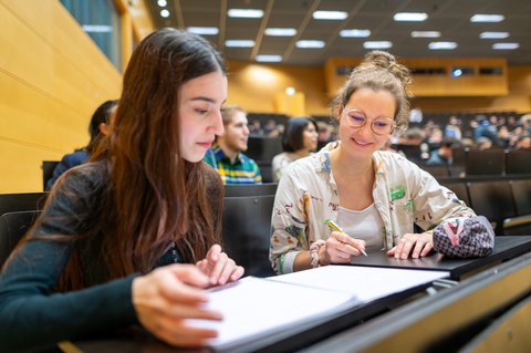 Zwei Studentinnen sitzen in einem Hörsaal, schreiben in ihre Hefte und tauschen sich aus. Im Hintergrund sind weitere Studierende zu sehen, die aufmerksam dem Vortrag folgen.