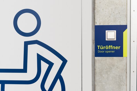 Rollstuhlsymbol auf einer Tür. Rechts davon der Schalter eines Türöffners.