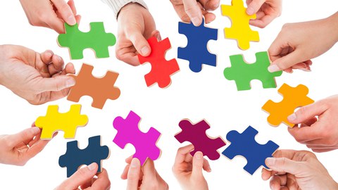 12 Hände ragen ins Bild. Jede Hand hält ein Puzzleteil. Jedes Puzzleteil hat eine andere Farbe.