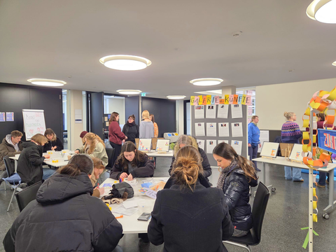 Studierende sitzen an Tischen und basteln. Im Hintergrund stehen Infotafeln und eine Ausstellung mit dem Titel „Galerie der Künfte“. Menschen tauschen sich in einer offenen und kreativen Atmosphäre aus.