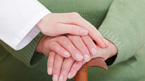 Auf dem Foto sind die Hände eines alten Mannes und einer jüngeren Frau zu sehen. Der ältere Mann hält einen Gehstock. Die jüngere Frau hat ihre Hand auf die des alten Mannes gelegt.