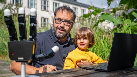 Das Foto zeigt einen Vater mit seinem Sohn. Beide sitzen an einem Tisch. Vor ihnen steht ein aufgeklappter Lptop sowie eine Roboterhand.
