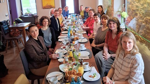 Eine Gruppe von Frauen sitzt an einem langen Tisch in einem Café beim Frühstück. Der Tisch ist reich gedeckt, die Stimmung wirkt freundlich. Die Umgebung ist gemütlich mit Steinwand und Deko.