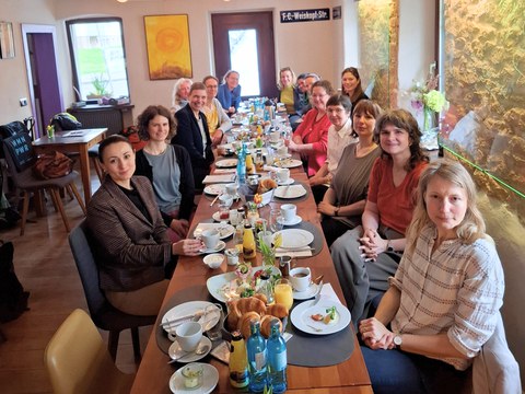 Eine Gruppe von Frauen sitzt an einem langen Tisch in einem Café beim Frühstück. Der Tisch ist reich gedeckt, die Stimmung wirkt freundlich. Die Umgebung ist gemütlich mit Steinwand und Deko.