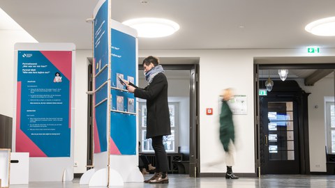 Person betrachtet Infotafeln in einer Ausstellung im Foyer, während andere vorbeigehen.