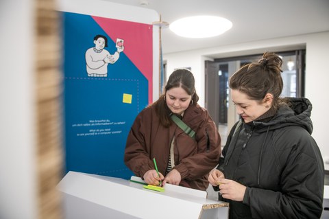Two people write notes on colorful slips at a white lectern in an exhibition in front of an info wall with an illustration.