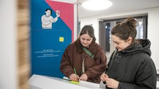 Two people write notes on colorful slips at a white lectern in an exhibition in front of an info wall with an illustration.