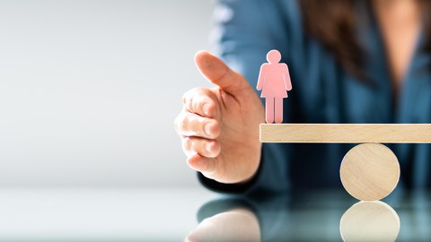 Foto einer Waage aus Holz. Auf einer Seite steht eine rosa Figur in weiblicher Form. Auf der anderen Seite eine blaue Figur in männlicher Form. Die Waage ist ausgeglichen. 