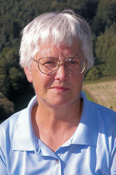 Portrait einer Frau mit kurzen weißen Haaren, Brille und einer hellblauen Bluse. Im Hintergrund ist Wald zu sehen.