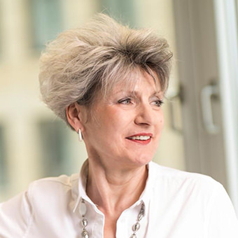 Das Bild zeigt eine Frau mit blonden kurzen Haaren. Sie trägt eine weiße Bluse und eine silberne große Kette. Sie schaut nach rechts und ist angeschnitten im Profil zu sehen.