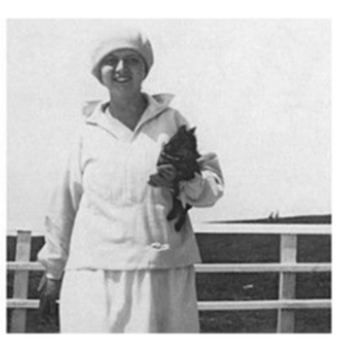It is a black and white photograph. A woman is standing on a railing. She is holding a black dog in her hand. She is wearing a light-coloured jumper and a white cap.