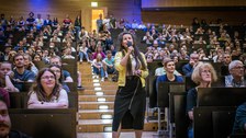 A woman is holding a microphone. She is wearing a yellow jacket and a black skirt. She is standing on a staircase in a lecture hall. To the left and right of her sits the audience.
