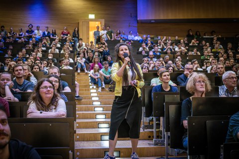 A woman is holding a microphone. She is wearing a yellow jacket and a black skirt. She is standing on a staircase in a lecture hall. To the left and right of her sits the audience.