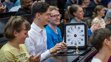 A man in a white shirt and dark hair is sitting at a table in the lecture hall. In front of him he has a large stopwatch with a black border.