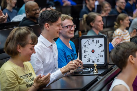A man in a white shirt and dark hair is sitting at a table in the lecture hall. In front of him he has a large stopwatch with a black border.