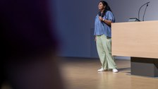 A woman is standing on a stage. She has black hair, a blue blouse, green trousers and white trainers. She holds a microphone in her hand and speaks to the audience.